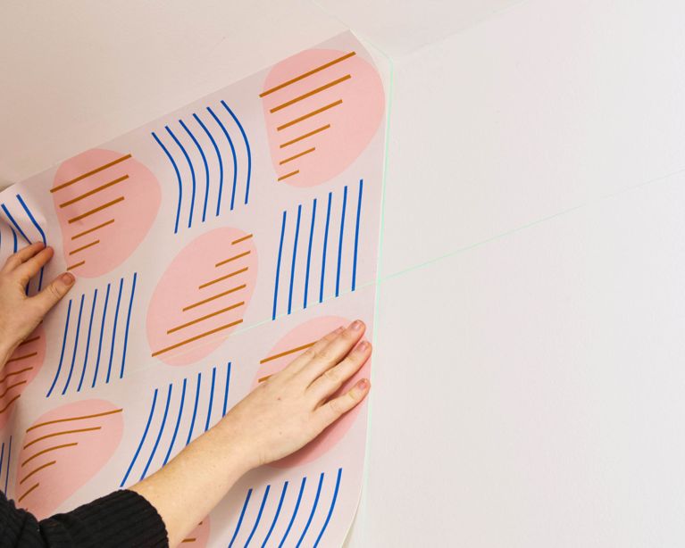 A woman installs the first panel of peel-and-stick wallpaper onto a white wall.
