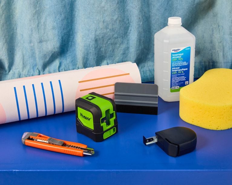 All of the tools needed to install peel-and-stick wallpaper: exacto knife, laser level, alcohol and sponge for cleaning, measuring tape and smoothing tool.