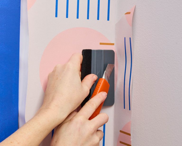 A woman trims the excess edge of peel-and-stick wallpaper with an exacto knife and straight edge.