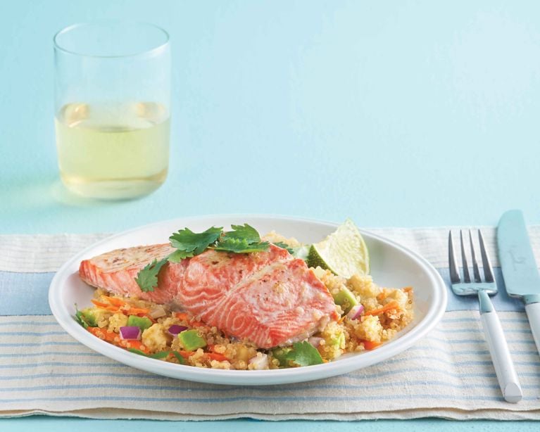 A bright, healthy meal featuring a flaky pink salmon fillet resting on a bed of fluffy quinoa. The salad is tossed with shredded carrots, diced red onion, and avocado. A lime wedge and fresh cilantro garnish the plate, set against a light blue background with a glass of white wine nearby.
