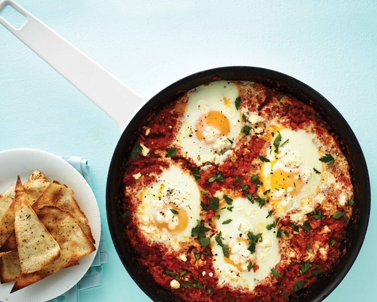 A black skillet filled with shakshuka, a thick red tomato and pepper sauce, with four eggs poached directly in the mixture. The eggs are garnished with crumbled white feta cheese and fresh chopped parsley. Beside the skillet is a white plate holding triangular, seasoned toasted pita chips, all set against a light blue background.