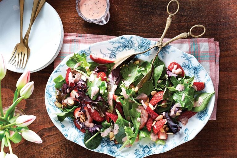 A top-down view of a spring salad served on a large, ornate blue floral platter. The salad features a mix of leafy greens, spinach, and purple radicchio, tossed with fresh strawberry slices and toasted almond slivers. A pink, creamy rhubarb vinaigrette with poppy seeds is drizzled over the top. Ornate brass serving tongs rest on the platter, and a small glass bowl of extra dressing sits nearby on a rustic wooden table next to pink tulips.