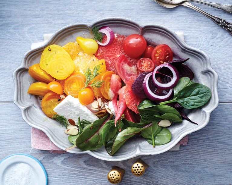 Overhead view of a vibrant spring superfood salad with yellow beets, tomatoes, red onions, and almonds, served in a vintage-style silver bowl on a light wood background.