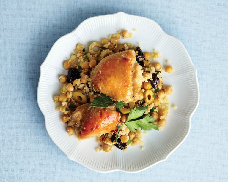 A top-down view of Moroccan-style sticky chicken thighs with a glossy glaze. The chicken sits atop a vibrant medley of fluffy couscous, whole chickpeas, sliced green olives, and dark dried plums (prunes), garnished with a fresh sprig of flat-leaf parsley.