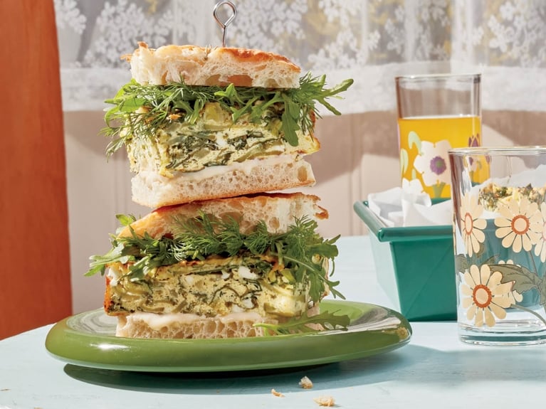 Two stacked sandwiches filled with green egg squares with feta.