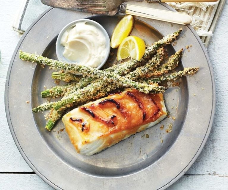 A grey plate with breaded asparagus stalks, a serving of fish, a side of white sauce and two lemon wedges.
