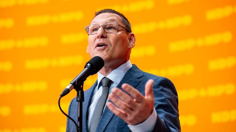 A photo of NDP federal leader Avi Lewis speaking at an event on an orange NDP background.