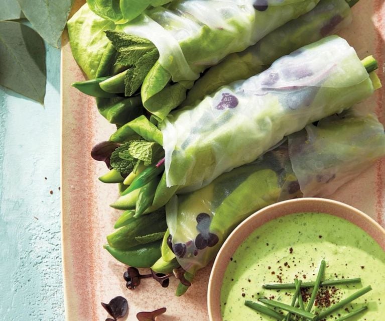 A closeup of rice-paper-wrapped green spring rolls and a small dish of green sauce topped with chives, on pink dishware