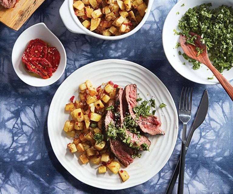 A white dish with sliced steak, cubed and fried potatoes and green sauce. Surrounding it are three white dishes containing a red sauce, more potatoes and more green sauce.