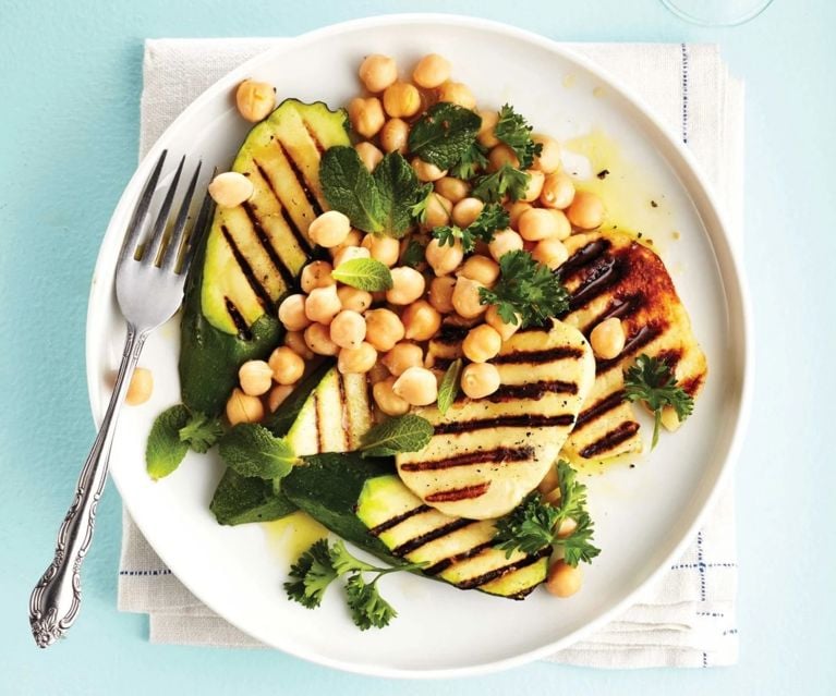 A white plate with a salad of chickpeas, halloumi, zucchini and other greens, as well as a fork. The plate sits on top of a folded white tea towel on a light blue surface.