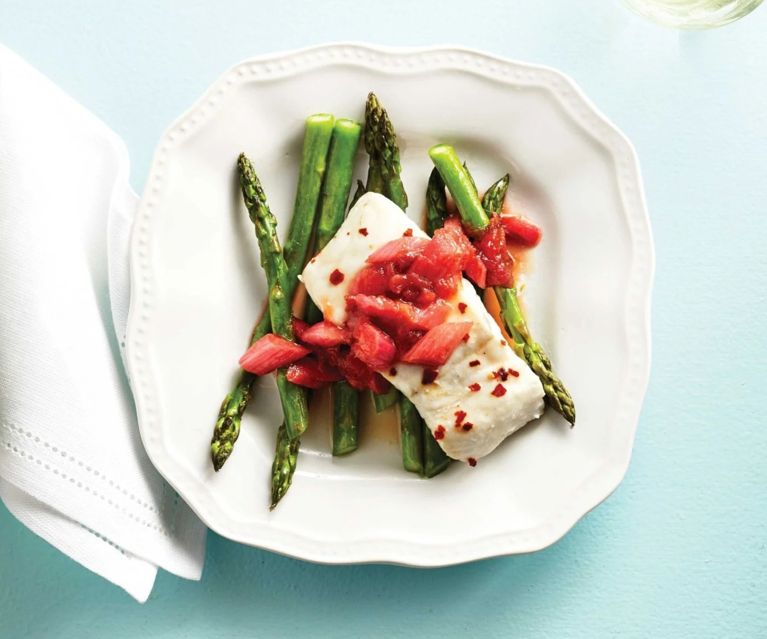 A serving of white fish topped with a rhubarb and bacon jam, resting on green asparagus and served on a white plate with wavy edges