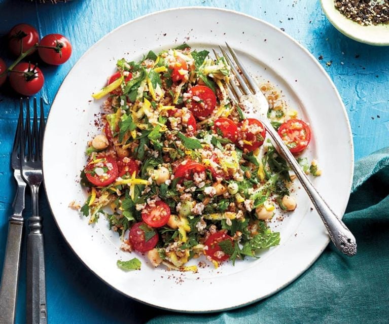 A white plate with tabbouleh with whole cherry tomatoes and fork on a bright blue surface, with a napkin, more forks and tomatoes visible around it