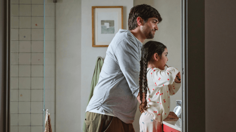 A father watches his young daughter brush her teeth.