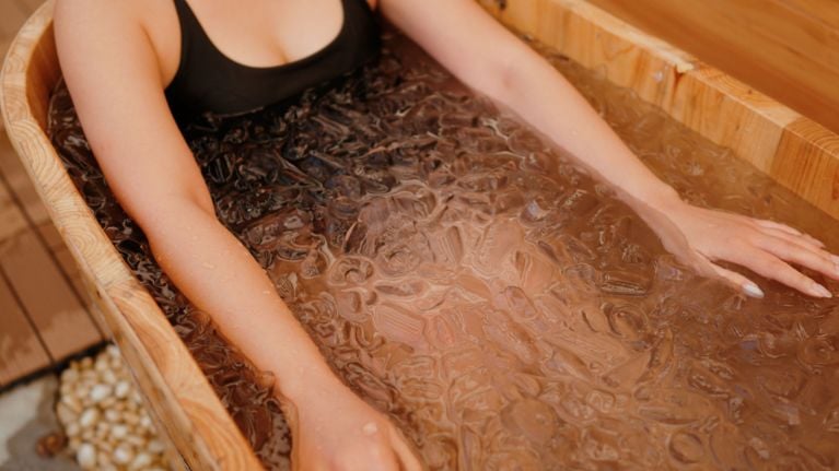 A woman takes an ice bath in a wooden tub, illustrating cold plunging.