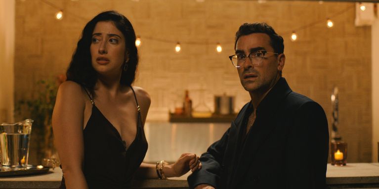 A photo of a woman in a black dress and a man in a black suit sitting at a bar used in a post about Dan Levy big Mistakes