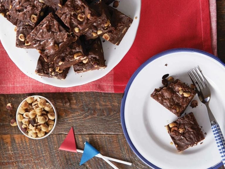 A blue-rimmed white plate of two hazelnut brownies with a blue and white fork resting on it next to a plate of more brownies and a bowl of hazelnuts and two small red and blue pennants for an article about the 18 best dessert square recipes