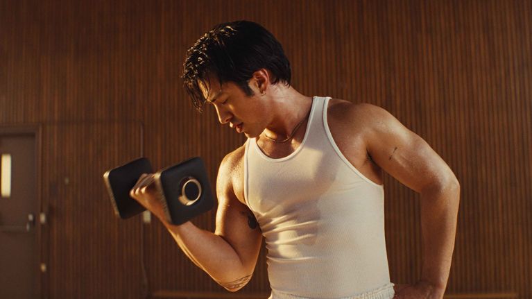 A man in a white tank top doing a bicep curl in a post about hudson williams peloton.