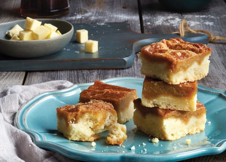 A stack of maple butter tart squares on a blue plate, next to a grey napkin with cubes of butter in a bowl on top of a cutting board in the background for an article about 17 Of Our Favourite Dessert Square Recipes To Make Now
