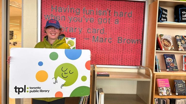 Author Marci Stepak holding a giant Toronto Public Library Card in front of a bulletin board that reads: Having fun isn’t hard when you’ve got a library card.