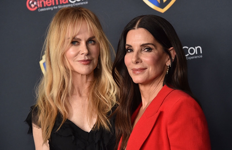 A photo of two women, one with long wavy red hair and one with long wavy brown hair, standing against a ComicCon logo wall in a post about Practical Magic 2.