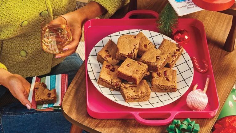 A person with a glass in hand, and a partially eaten Pumpkin Brown Butter Blondie in the other. A plate of blondies is set on a tray beside them for an article about the 18 best dessert square recipes