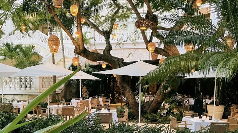 Tables with white table cloths and umbrellas on a patio surrounded by palm trees and hanging rattan light shades in a post about rosewood baha mar review.