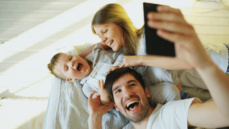 Smiling parents with baby taking selfie family photo on bed at home.