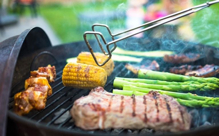 A pair of silver tongs suspended over an open black grill with various meats and vegetables grilling.