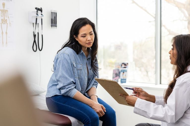 A woman sitting on a stretcher in.a doctor's office looking at paperwork from her doctor in a post about PMOS