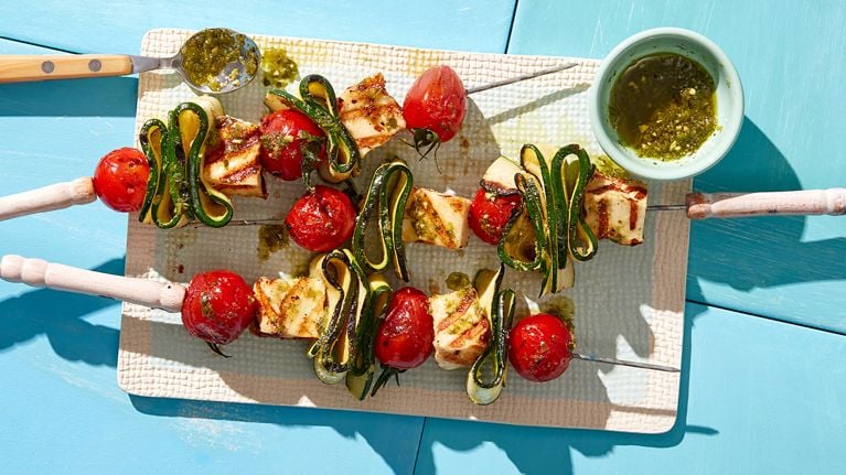 Three tomato, zucchini and halloumi skewers on a board with pesto sauce.