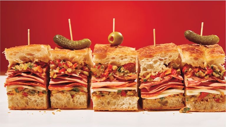 A giant muffaletta sandwich sliced into three pieces, with a pickle and olive on top as garnish.