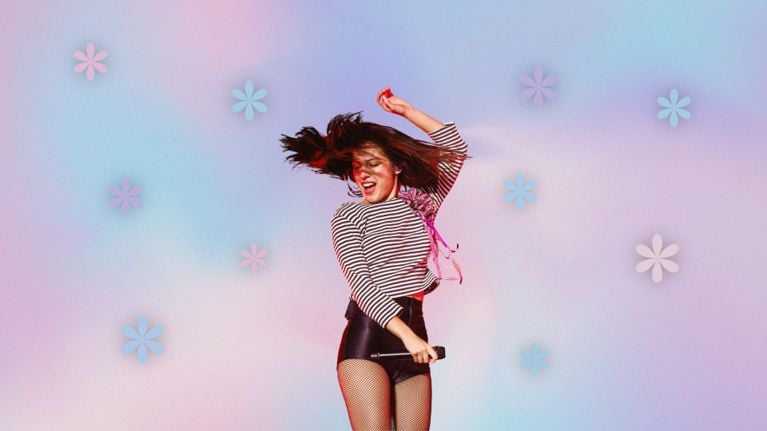 Olivia Rodrigo dances while holding a microphone, on a multi-coloured pastel background with colourful flowers around her