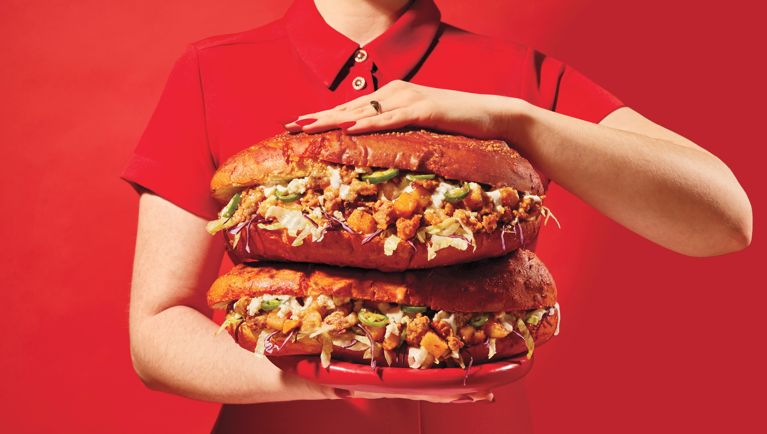 A woman in a red shirt holding two giant Pambazzo De Chorizo (Salsa-Dunked Chorizo Sandwiches) on a red background