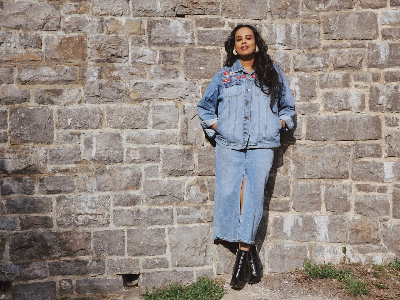 A woman, Meseret Haileyesus, wearing a denim jacket and skirt, stands by a rock wall looking pensively ahead of her, for a story about financial abuse in Canada.