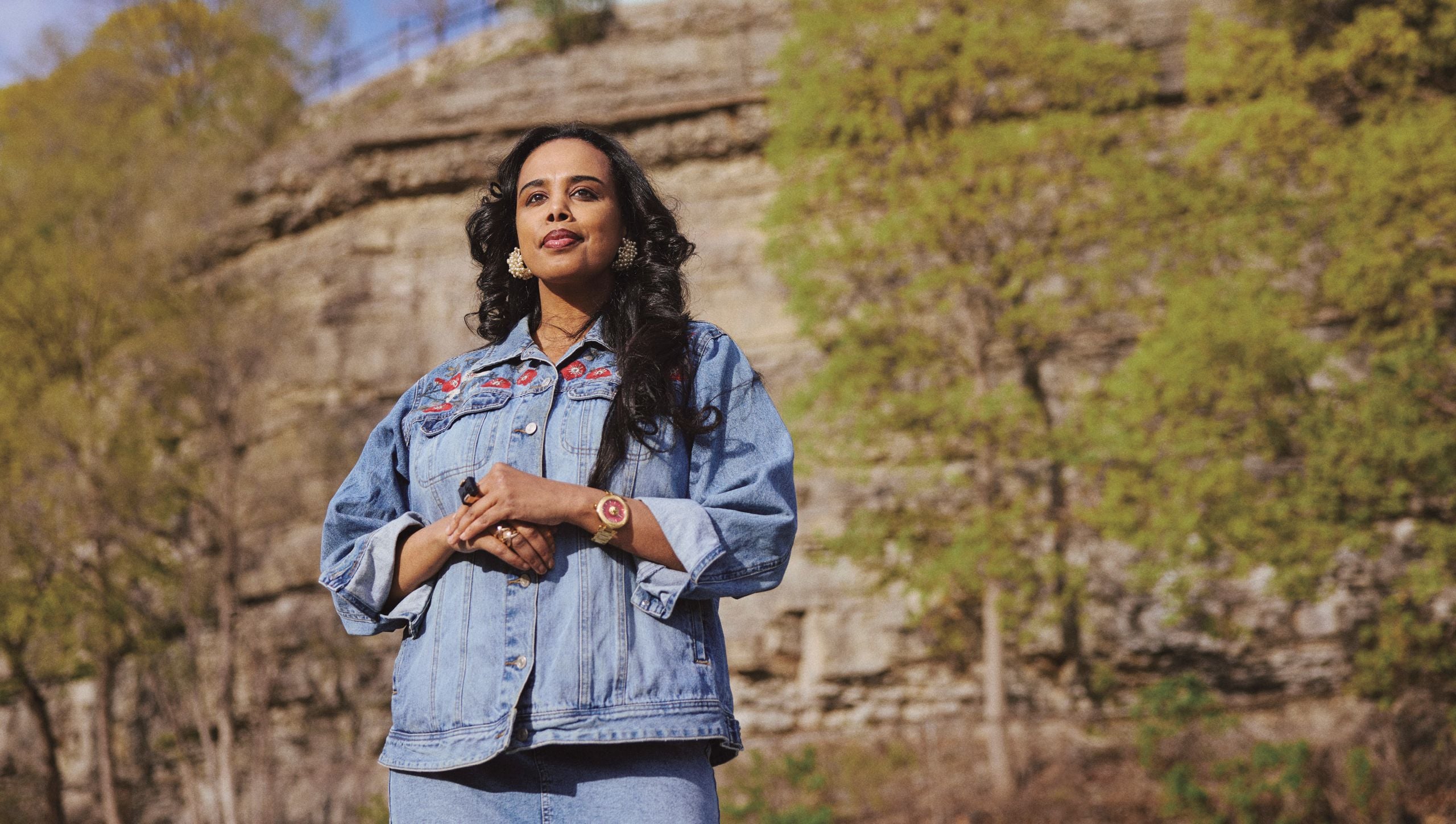 A woman, Meseret Haileyesus, wearing a denim jacket and skirt, stands by a rock wall looking pensively ahead of her, for a story about financial abuse in Canada.