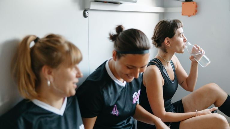 Three women sitting in black sport uniform, with girl on the far right drinking out of a water bottle